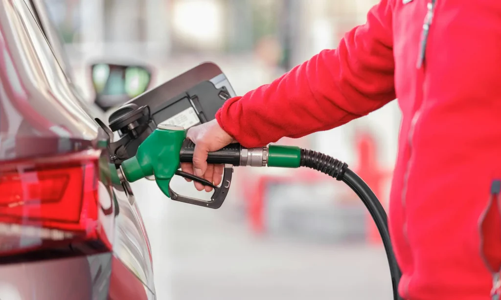 Aide carburant : le gouvernement élargit le dispositif des « grands rouleurs » à 5,5 millions de Français Un homme habillé en polaire rouge, fait le plein d'essence d'une voiture avec un pistolet de pompe a essence vert.